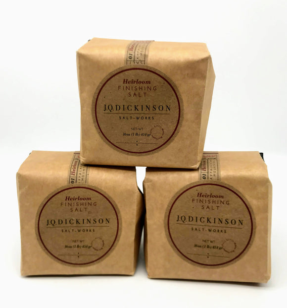 Three small brown paper bags (sealed and filled with salt) are stacked upon one another against a plain white background. The labels read JQ Dickinson Salt Works Heirloom Finishing Salt.