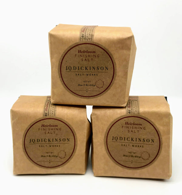 Three small brown paper bags (sealed and filled with salt) are stacked upon one another against a plain white background. The labels read JQ Dickinson Salt Works Heirloom Finishing Salt.